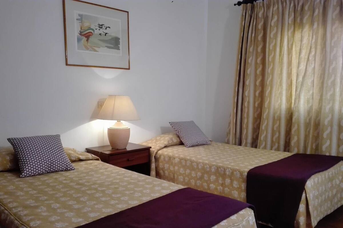 A room with two beds, each with a patterned bedspread and a purple blanket. There is a nightstand with a lamp between the beds, and a framed picture on the wall.