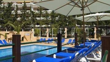 A swimming pool with blue lounge chairs and umbrellas, surrounded by palm trees and buildings.