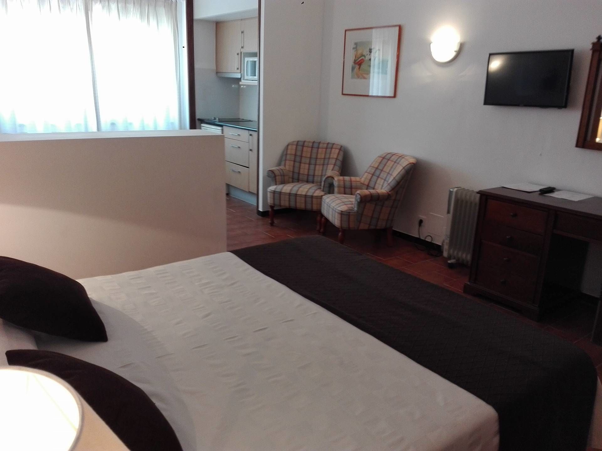 Hotellihuoneessa on sänky, tuoli, televisio ja taustalla näkyy pieni keittiönurkkaus.