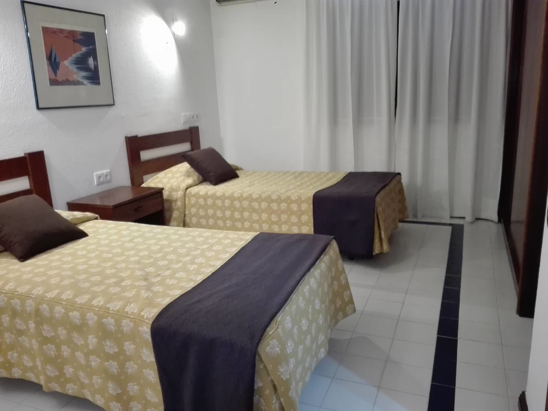 A hotel room with two beds, each with a patterned bedspread and a dark-colored blanket. There is a nightstand with a lamp, a wooden headboard, and a framed picture on the wall.