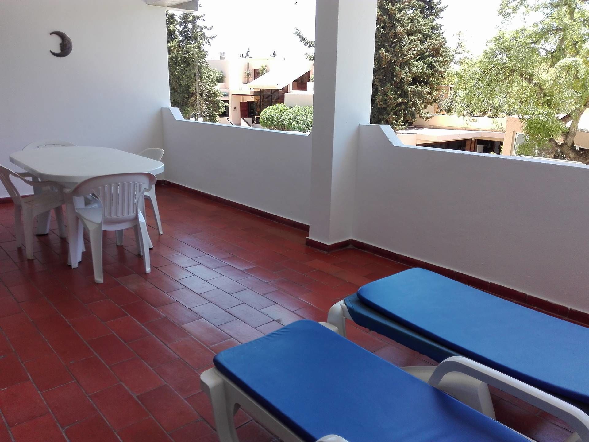 A balcony with a red-tiled floor, white walls, and a view of buildings and trees. There are two blue cushioned seats and a white table with chairs.