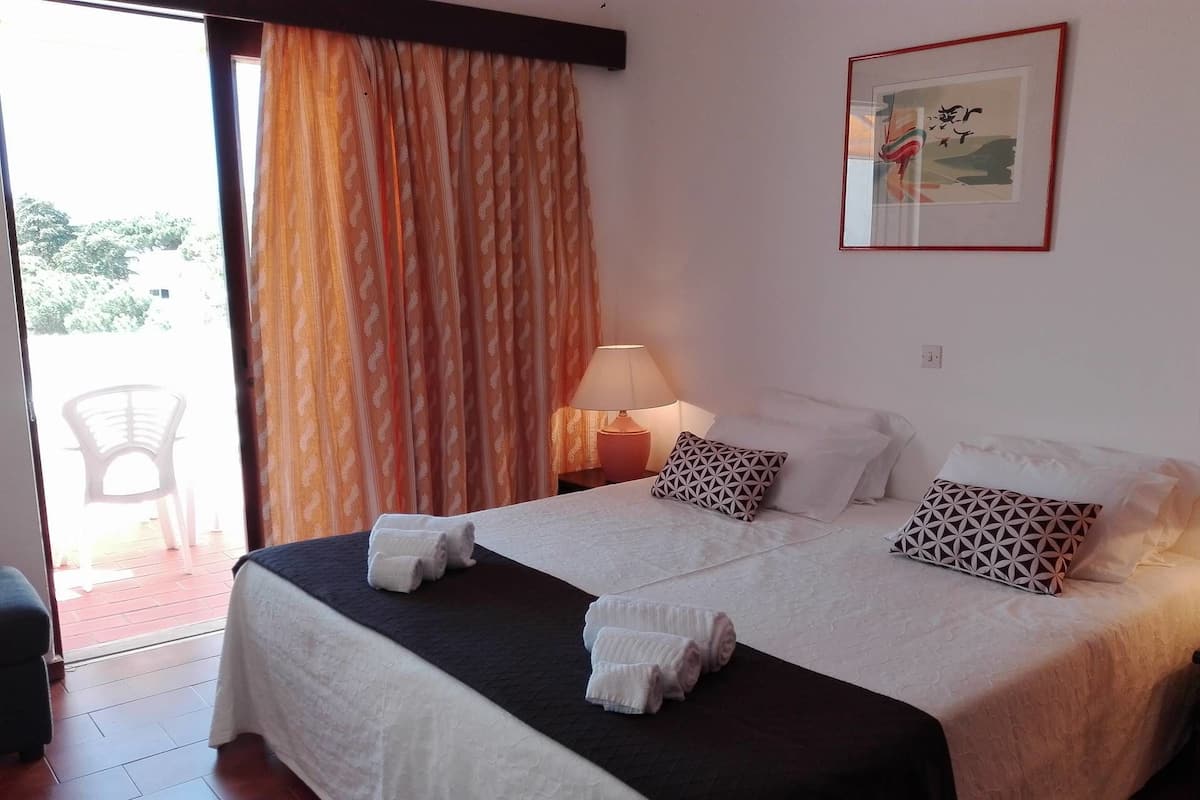 A double bed with a black and white bedspread, two pillows, and two rolled towels. There is a nightstand with a lamp and a framed picture on the wall.