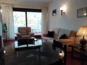 A living room with a sofa, armchair, and coffee table. There is a TV on a stand, a lamp, and a window with curtains.