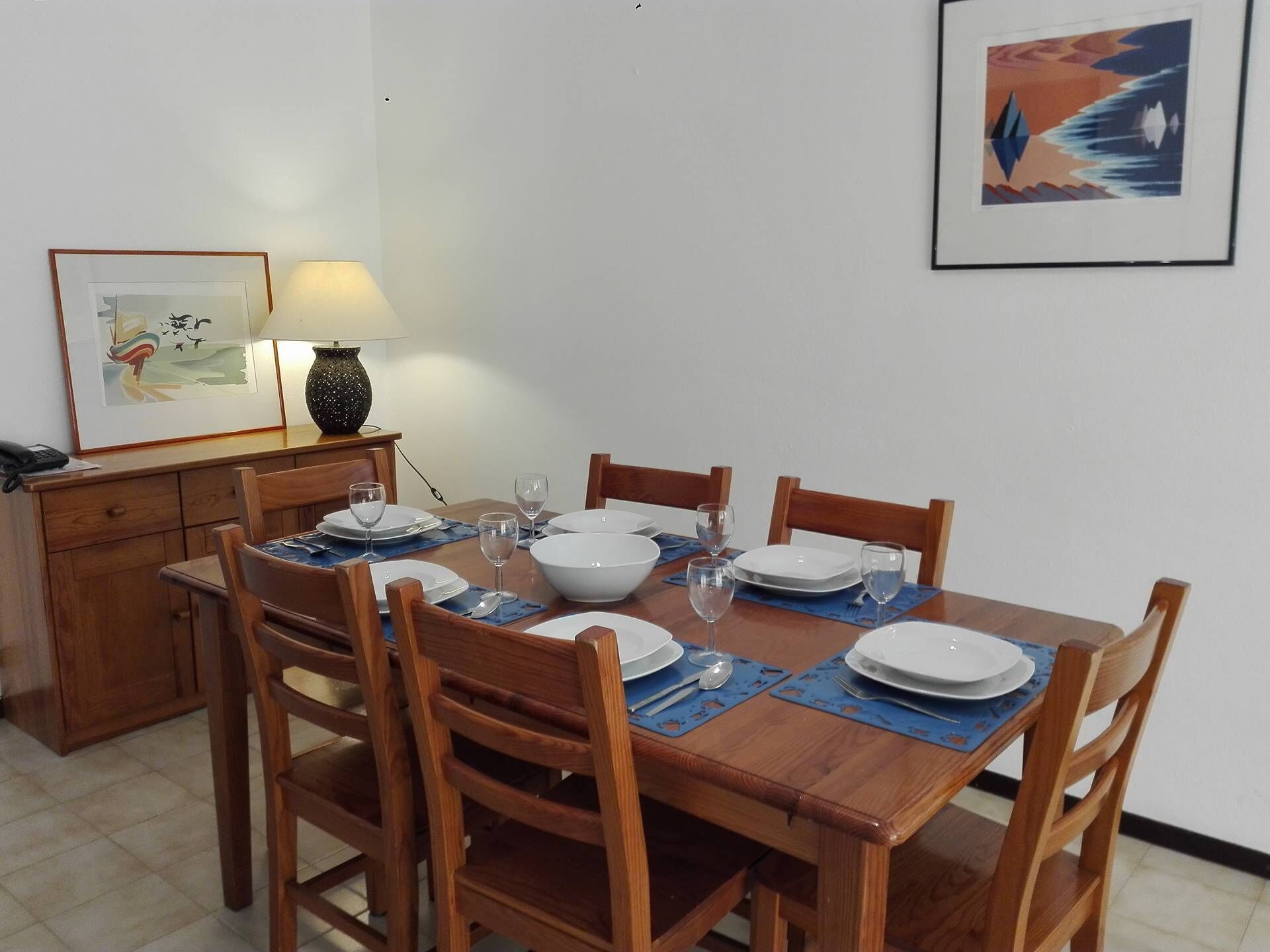 Un comedor con una mesa de madera dispuesta para seis personas, rodeada de sillas de madera. Hay un florero en la mesa, y un cuadro enmarcado cuelga de la pared a la izquierda.