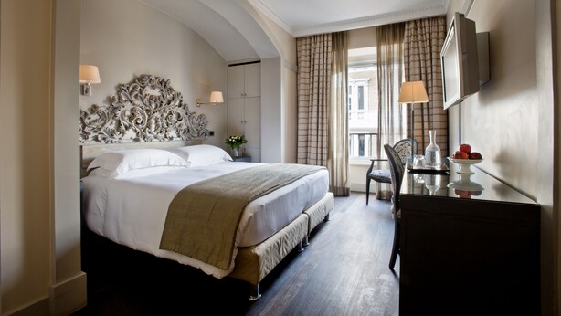 A hotel room with a large bed, a desk with a lamp, a television, and a view of the outside through the window. - Casa Montani (Rome)