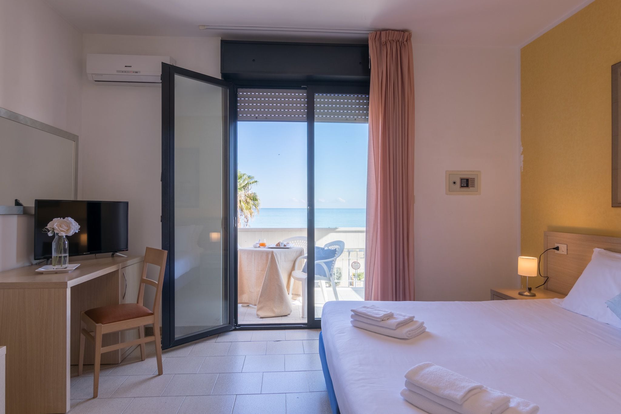 A hotel room with a bed, a desk with a chair, a television, and a balcony with a table and chairs.