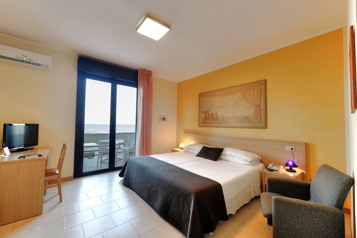 Ein Hotelzimmer mit Bett, Schreibtisch, Stuhl und Meerblick durch ein großes Fenster.