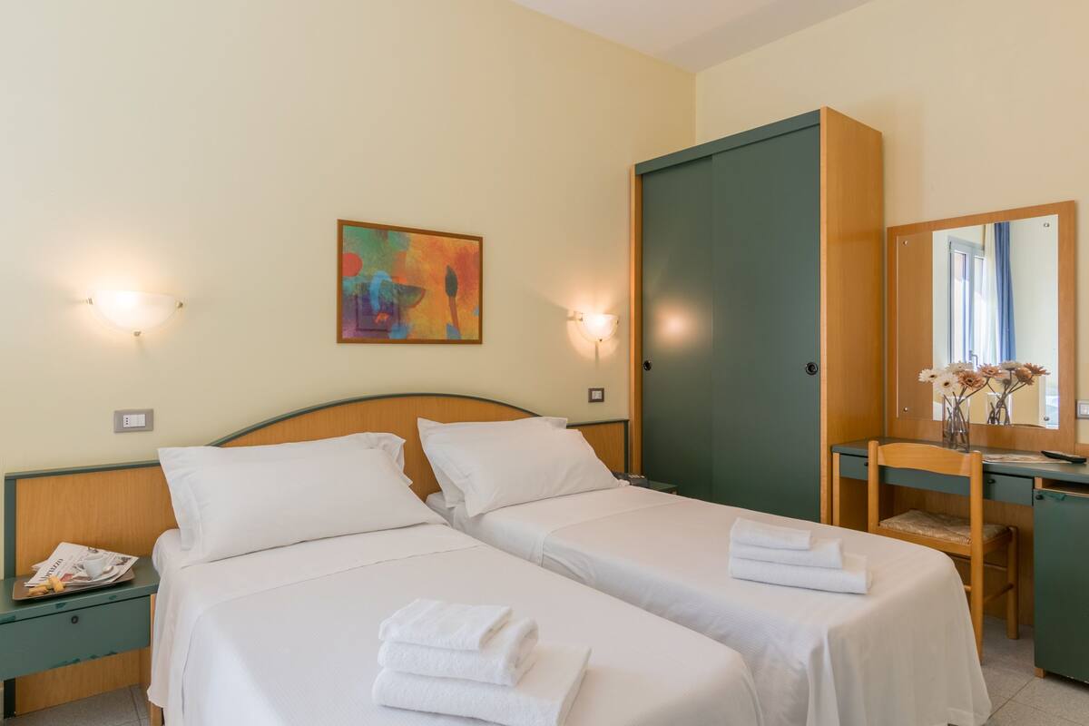 Ein Hotelzimmer mit zwei Betten, einem Schreibtisch, einem Stuhl, einem Spiegel und einem Gemälde an der Wand.