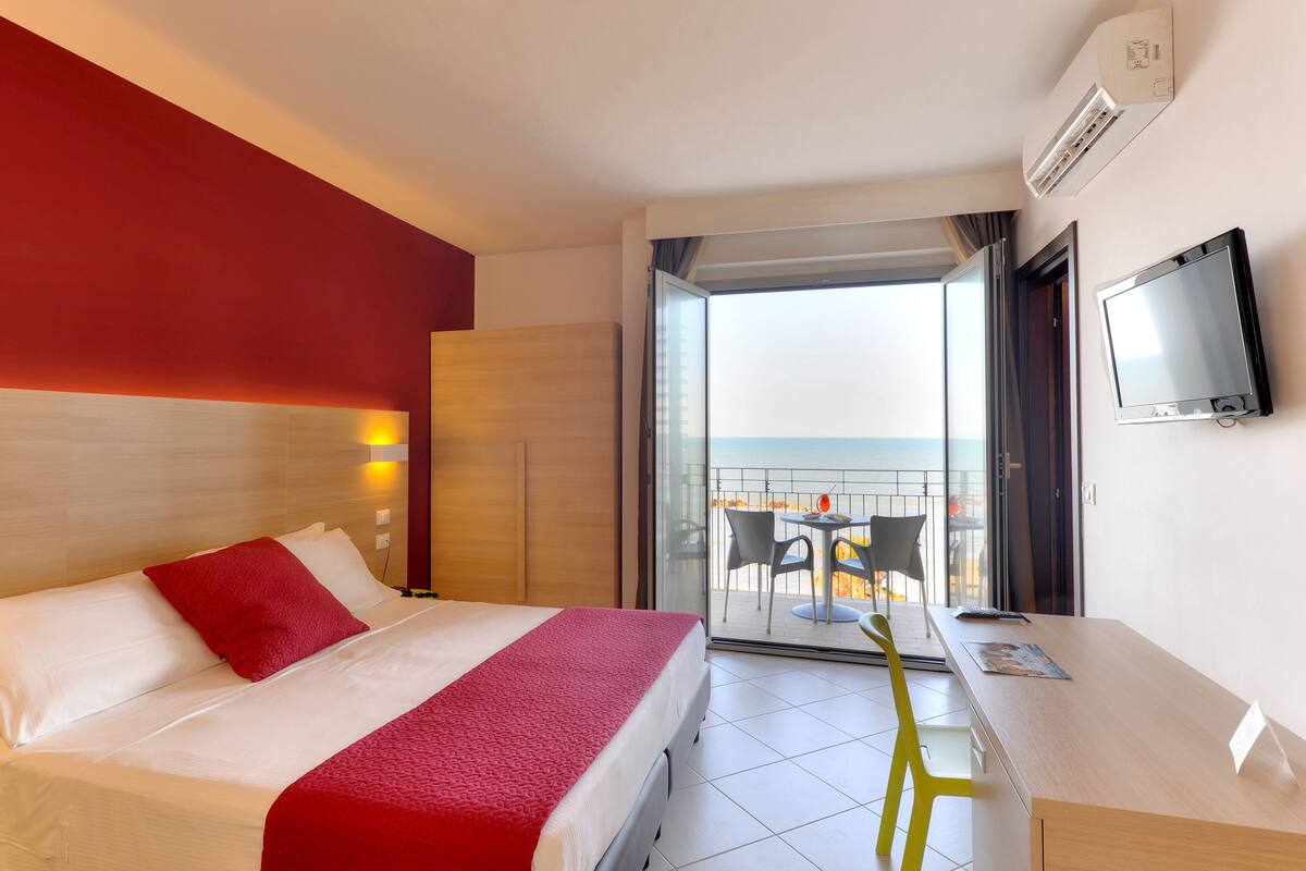 Ein Hotelzimmer mit Bett, Schreibtisch, Fernseher, einem Balkon mit Tisch und Stühlen sowie Meerblick.
