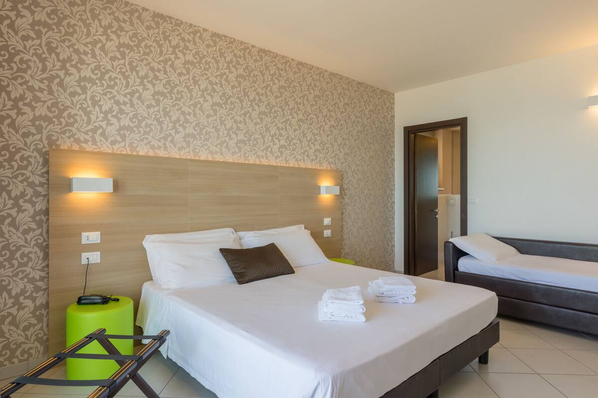 Ein Hotelzimmer mit einem großen Bett, einem kleineren Bett, einem grünen Hocker und einer Leiter.