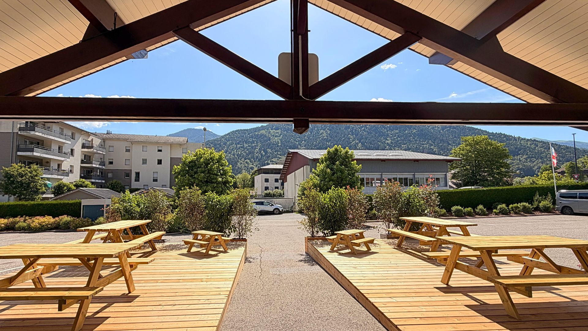 Zona d'estar a l'aire lliure coberta, amb taules de picnic i bancs de fusta, amb vista a un aparcament i edificis amb un paisatge de muntanyes al fons.