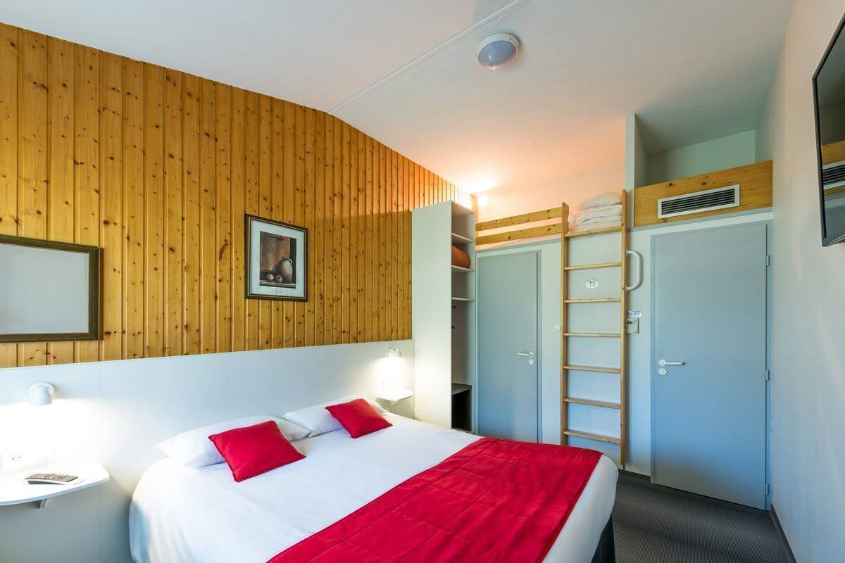 A hotel room with a wooden wall panel, a bed with red pillows, a bedside table with a lamp, and a wardrobe with a ladder.