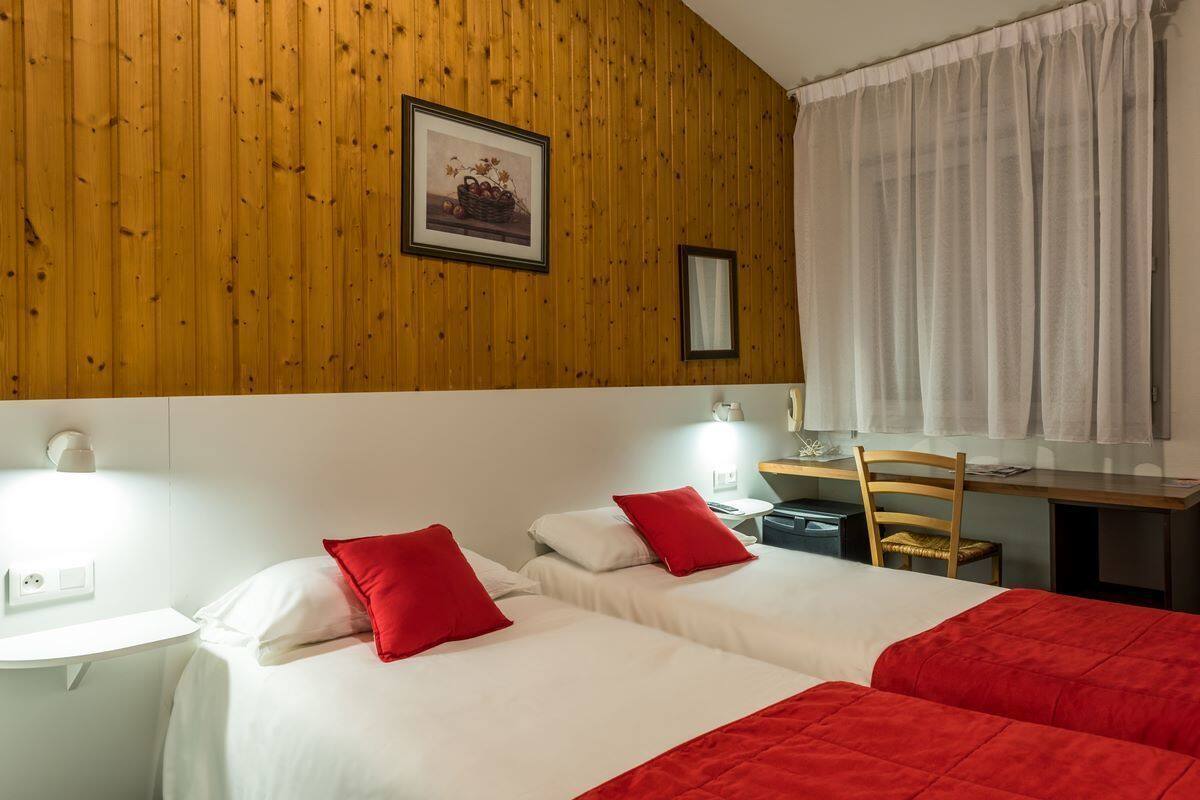 A hotel room with two beds, a desk, and a chair. There is a framed picture on the wall and a window with curtains.