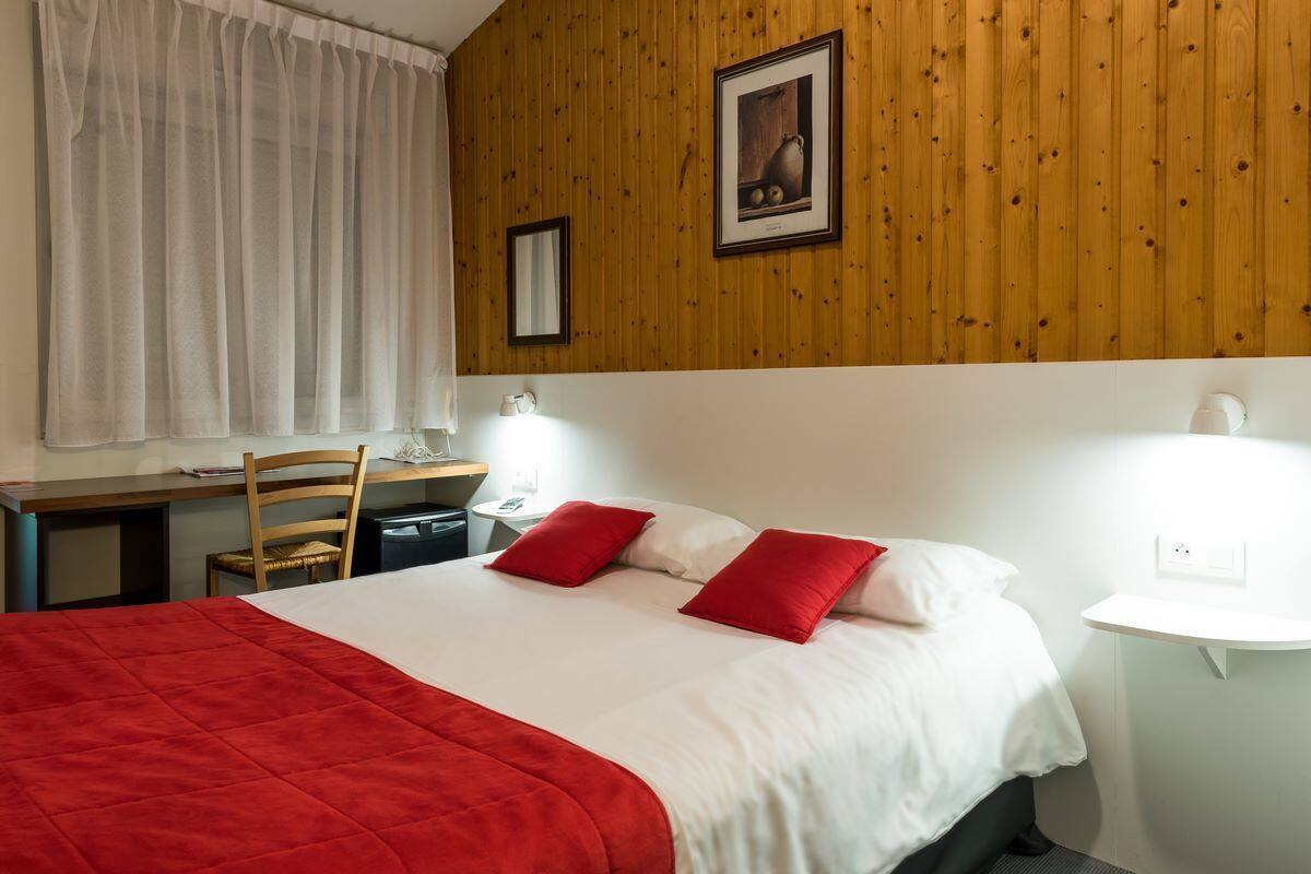 A hotel room with a wooden ceiling, a bed with red and white bedding, a desk with a chair, a small bedside table with a lamp, and a window with white curtains.