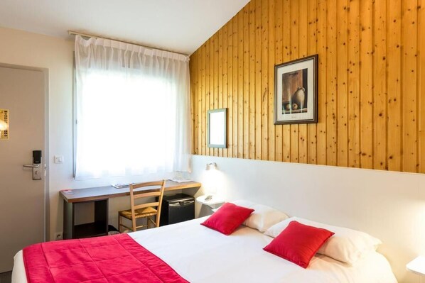 Habitación de hotel con pared de paneles de madera, cama con ropa de cama roja y blanca, escritorio con silla y ventana con cortinas blancas.