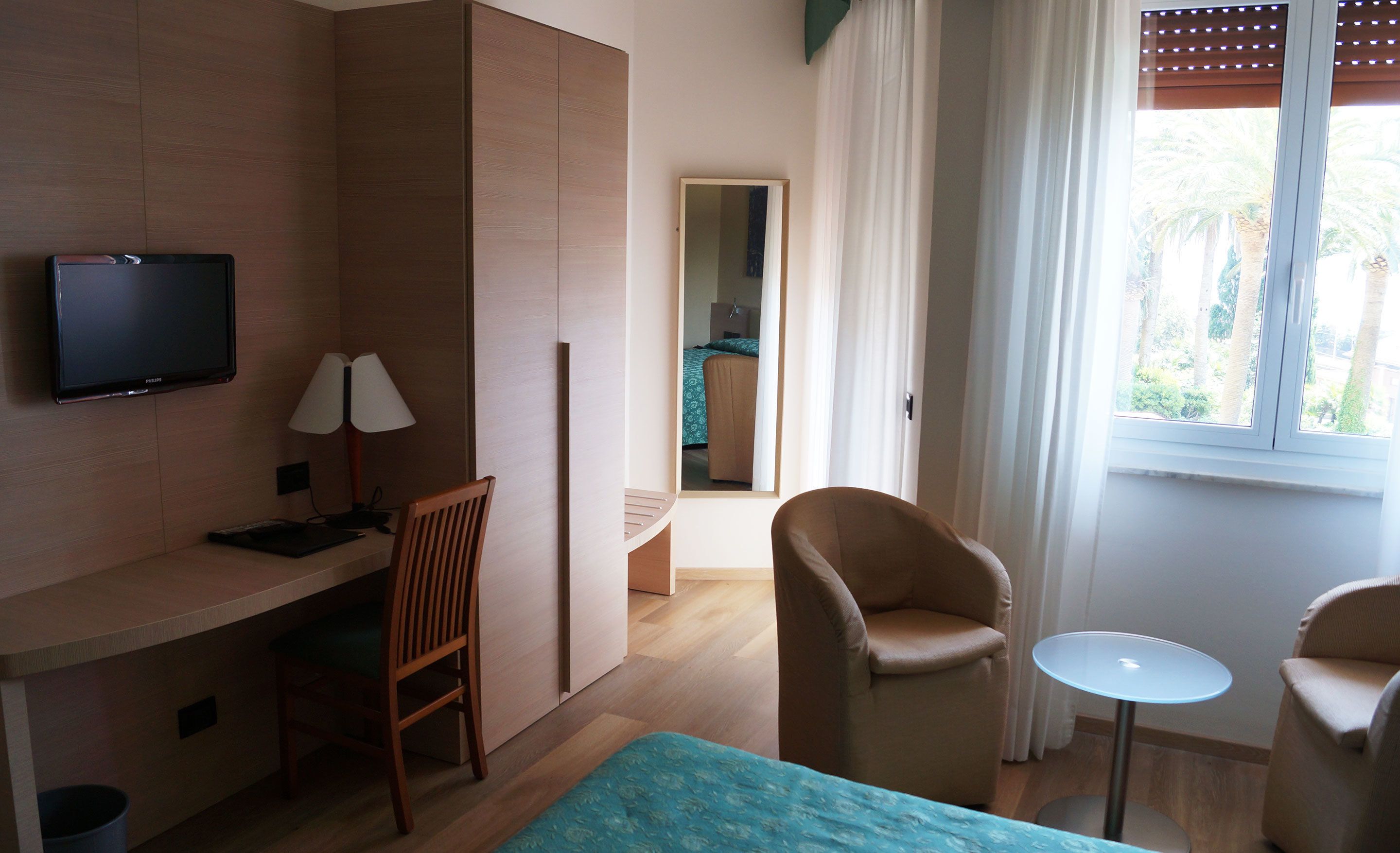 Ett hotellrum med en säng, ett skrivbord med en dator, en stol, en tv och ett fönster med gardiner.