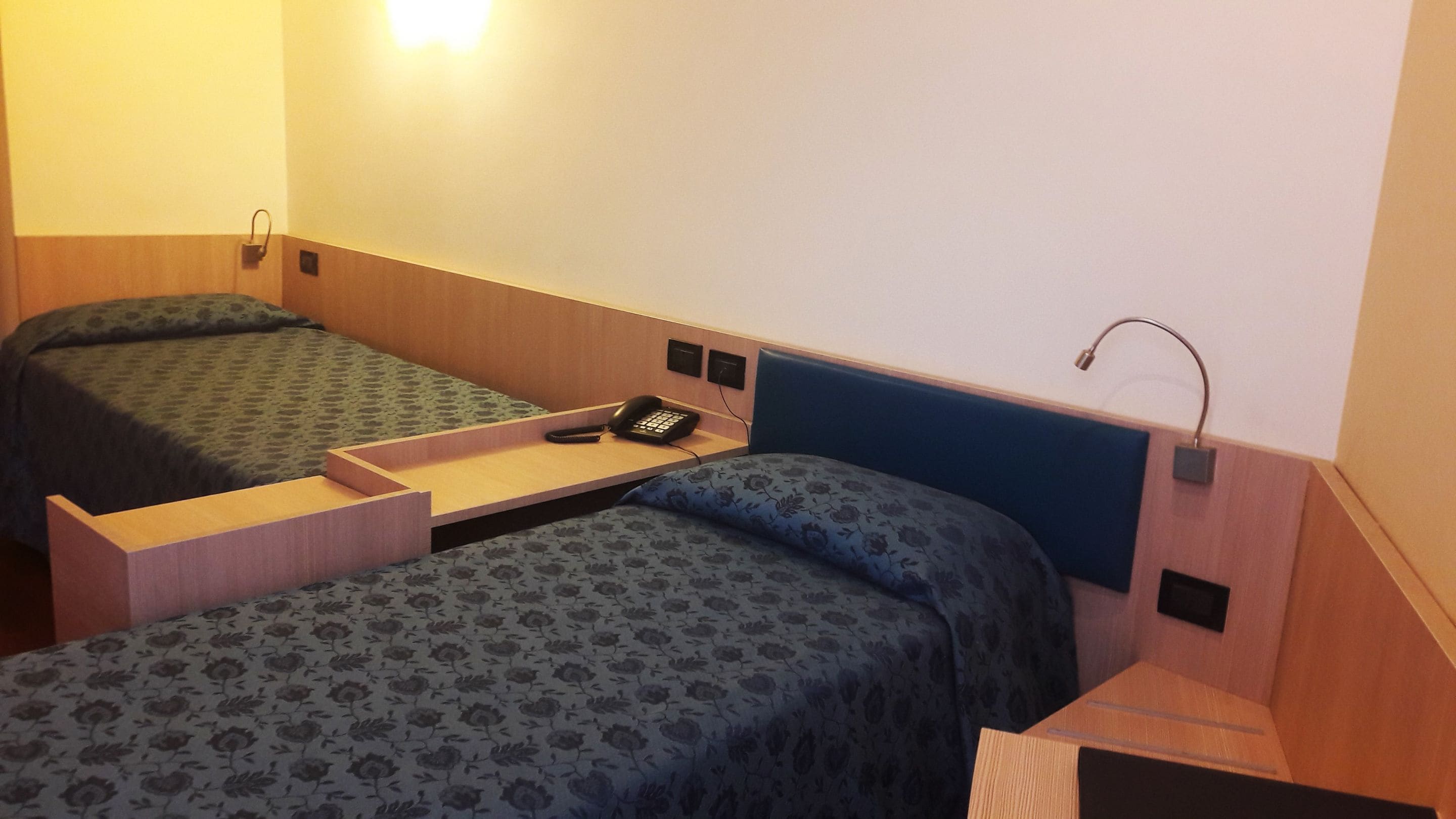 A small, compact room with two single beds, a bedside table with a phone, and a wall-mounted lamp.