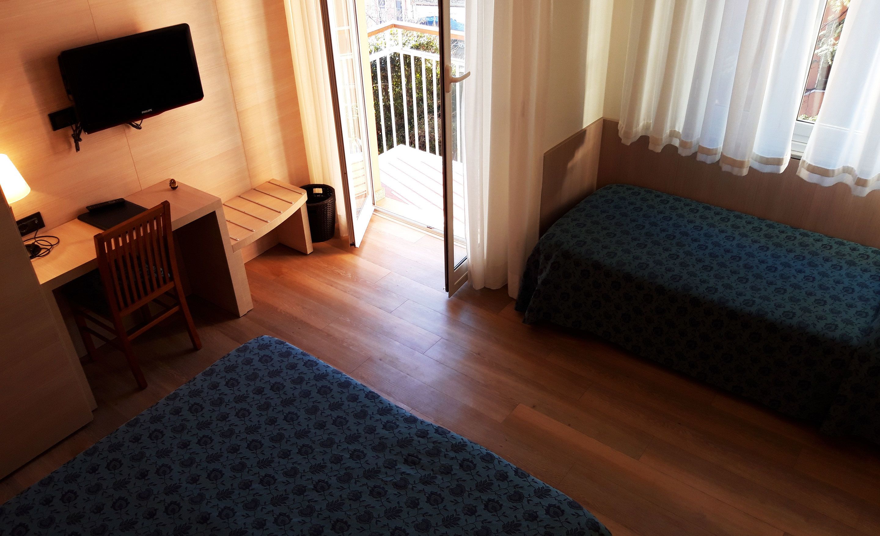 Ein kleiner, sauberer Raum mit zwei Einzelbetten, einem Schreibtisch mit Stuhl, einem Fernseher und einer Glastür zum Balkon.
