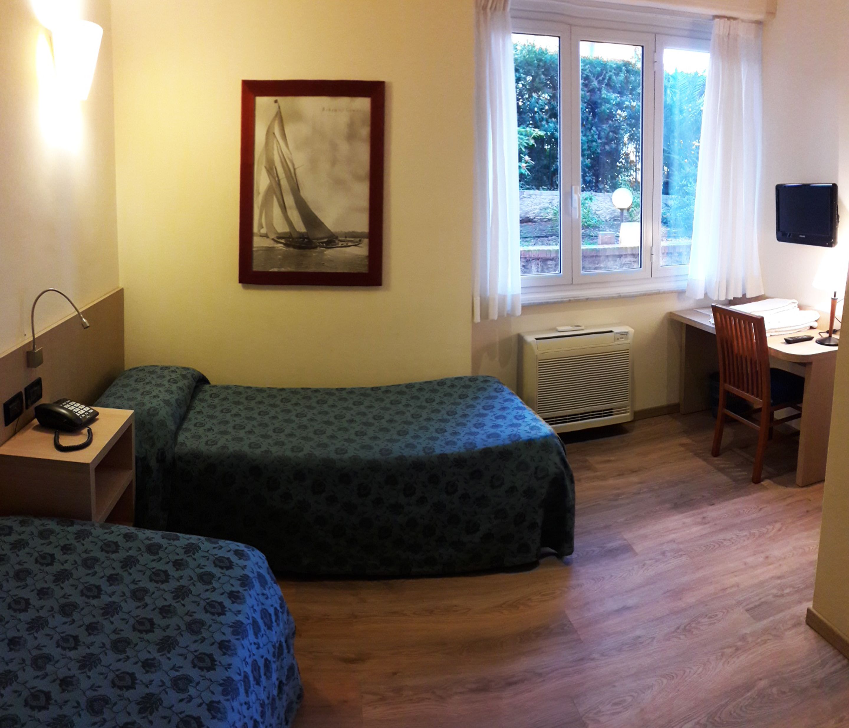 Habitación de hotel con dos camas, un escritorio, una silla, un radiador, un televisor y una ventana con vistas a zonas verdes.