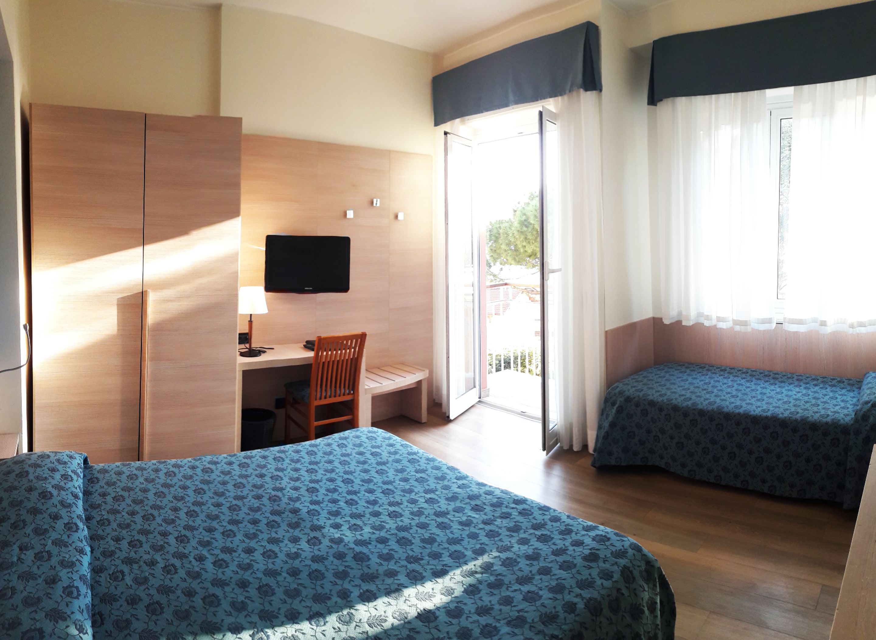 A hotel room with two beds, a desk with a chair, a television, and a sliding door to a balcony.
