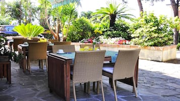 An outdoor patio area with wicker furniture, stone flooring, and a variety of plants and flowers.