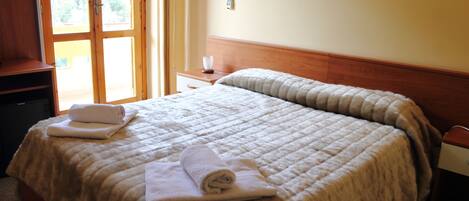 Habitación de hotel con una cama grande, un cabecero de madera, una mesita de noche, un escritorio y una ventana con vistas al verdor exterior.