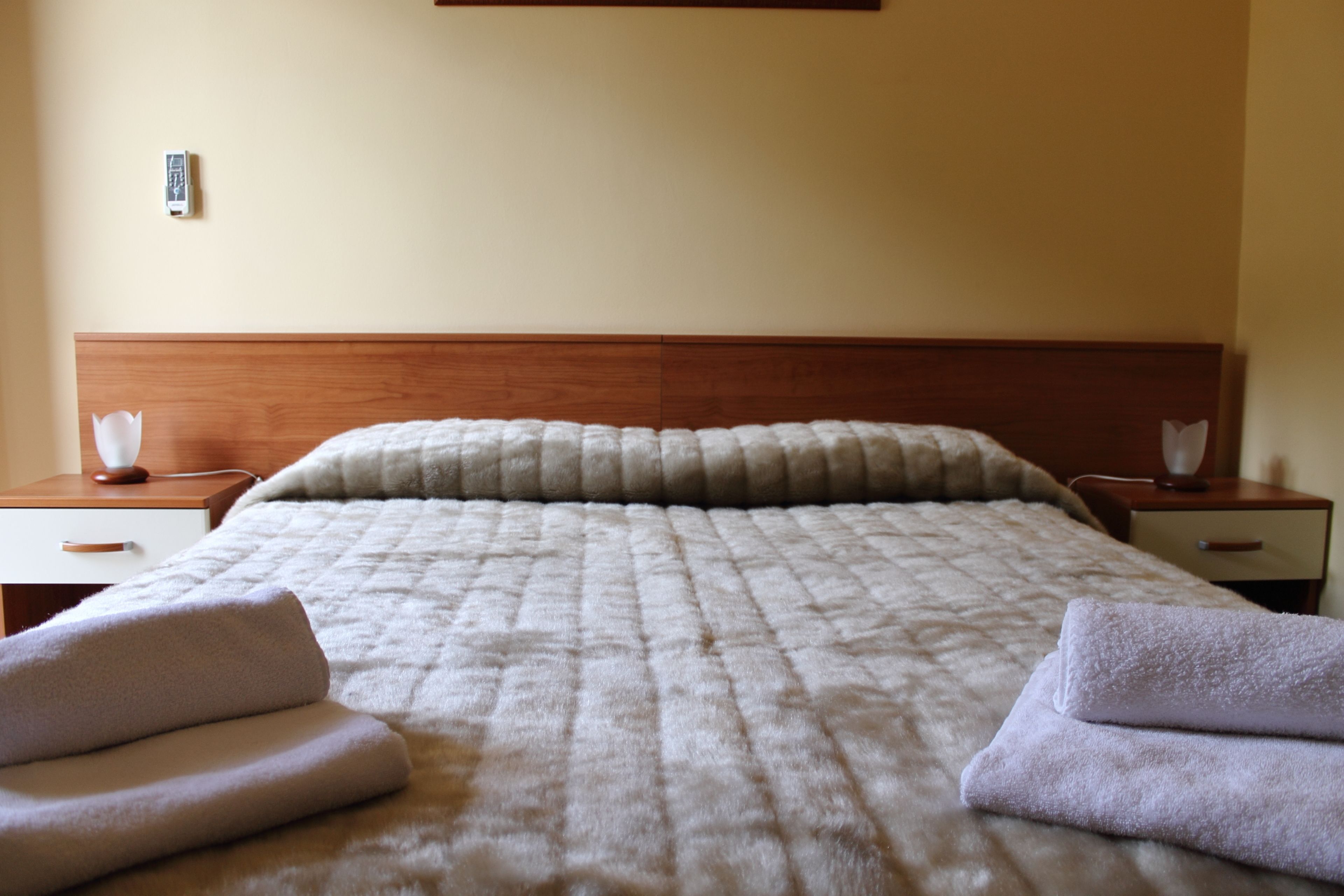 Ein ordentlich bezogenes Bett mit einer gesteppten Decke, zwei Nachttische mit Lampen und ein an der Wand befestigtes Thermostat.