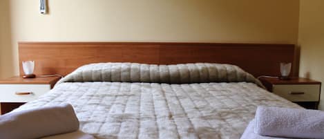 Cama bem arrumada com manta acolchoada, duas mesas de cabeceira com abajures e um termostato fixado na parede.