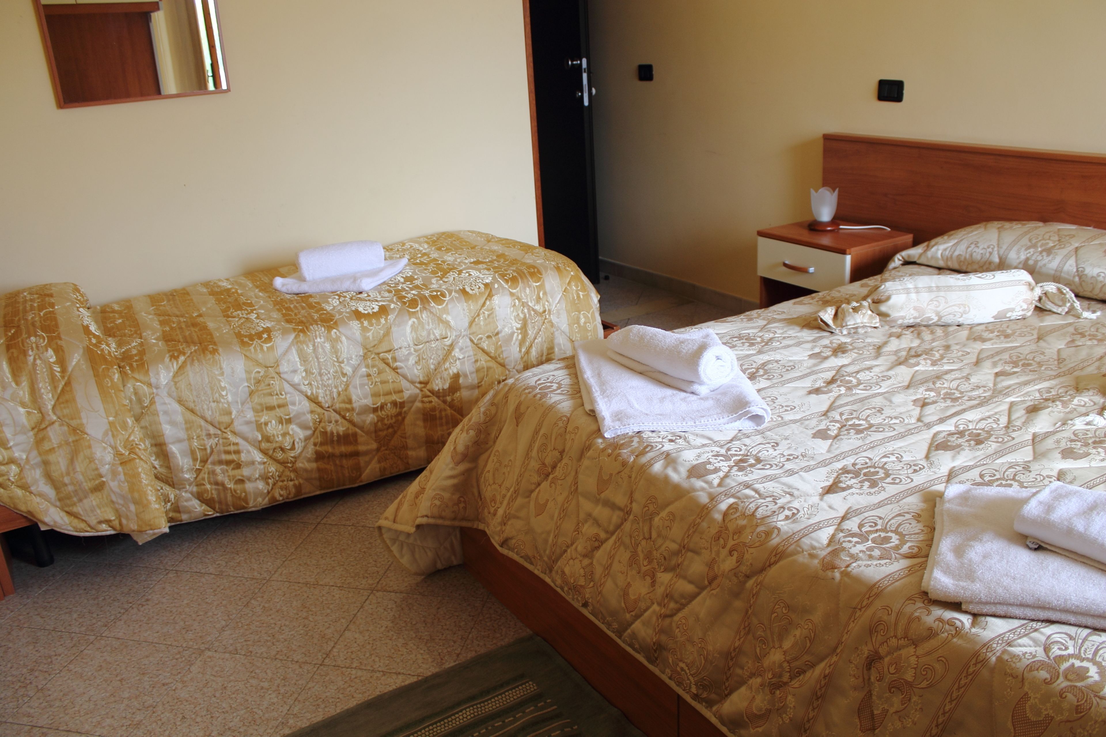 Ein Hotelzimmer mit zwei Betten, jeweils mit einem gemusterten Überwurf und weißen Handtüchern. Ein Nachttisch mit Lampe und einer kleinen Schublade ist vorhanden.