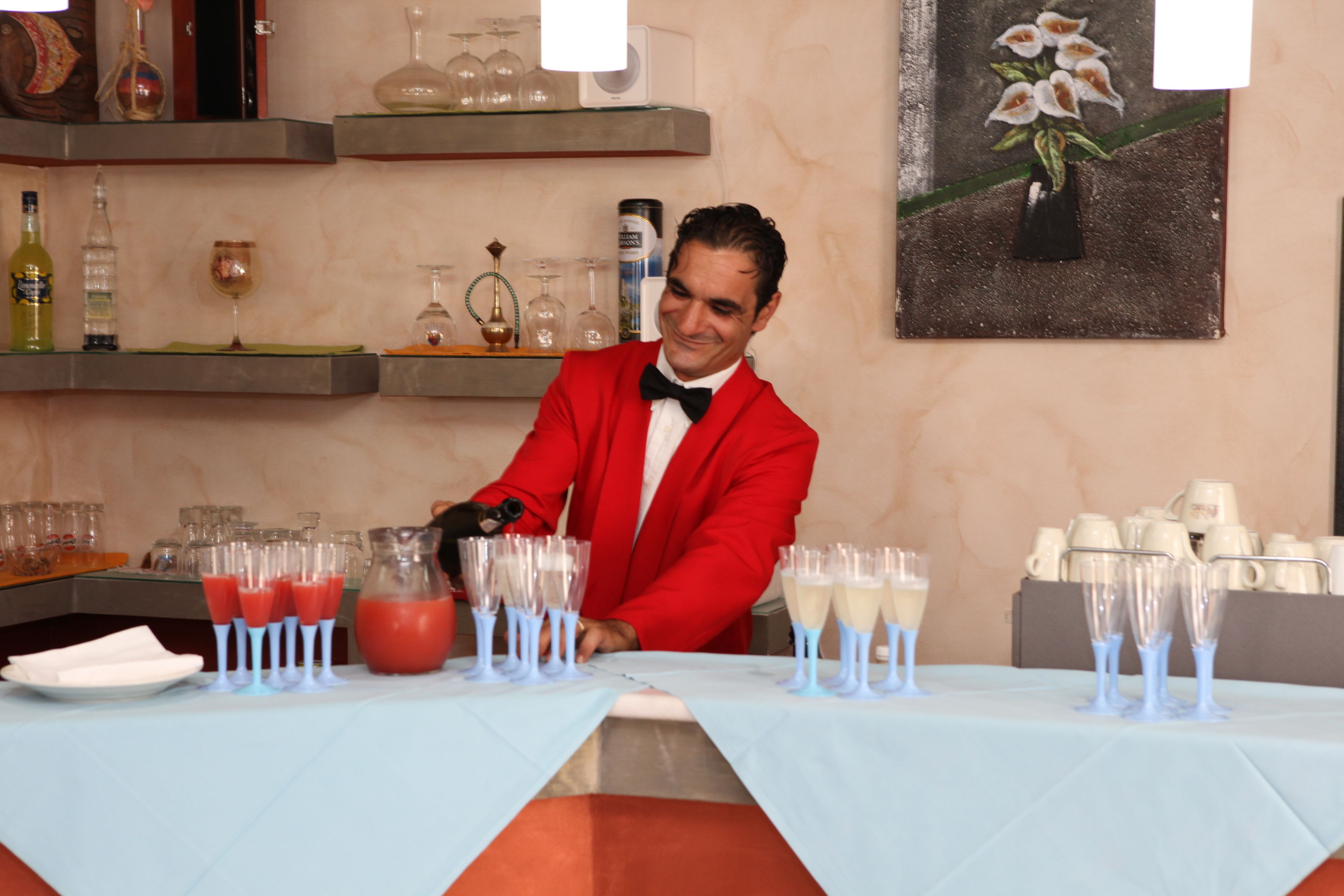 A bar area with a bartender in a red suit, glasses, and bottles on shelves.