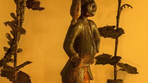 Una estatua de una persona con cabeza y cuerpo detallados, rodeada de plantas de hojas grandes.