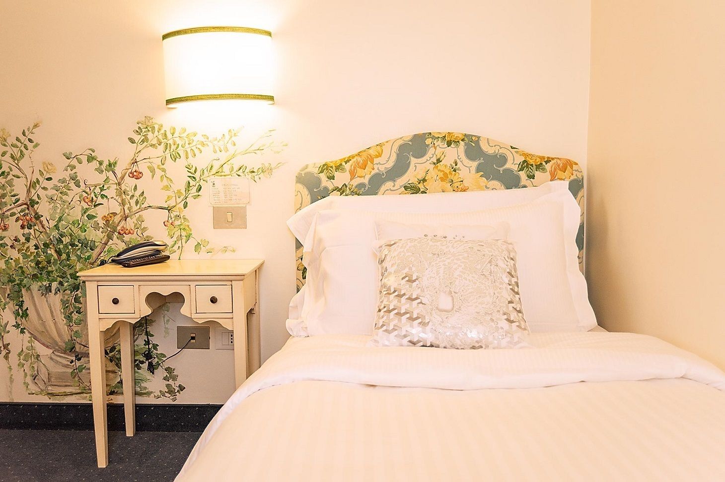 A neatly made bed with a floral headboard, a bedside table with a lamp, and a wall with a floral wallpaper design.