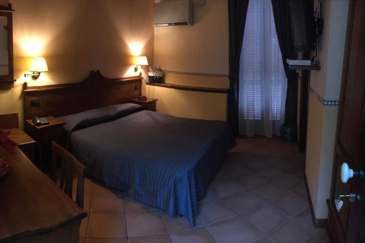 A small, dimly lit hotel room with a single bed, a wooden nightstand, a desk with a lamp, and a mirror.