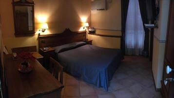 Una habitación de hotel pequeña y con poca luz, con una cama individual, una mesita de noche de madera, un escritorio con una lámpara y un espejo.