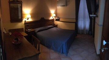 Een kleine, zwak verlichte hotelkamer met een enkel bed, een houten nachtkastje, een bureau met een lamp en een spiegel.