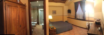 Una habitación con un armario de madera, una cama con manta azul, una mesita y un televisor de pared. Se ve un baño a través de una puerta abierta.