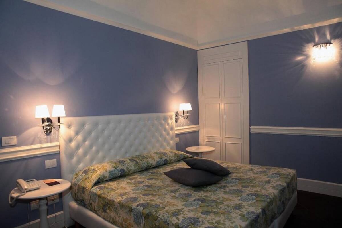 A bedroom with a tufted headboard, a floral bedspread, two bedside lamps, a bedside telephone, and a round table.