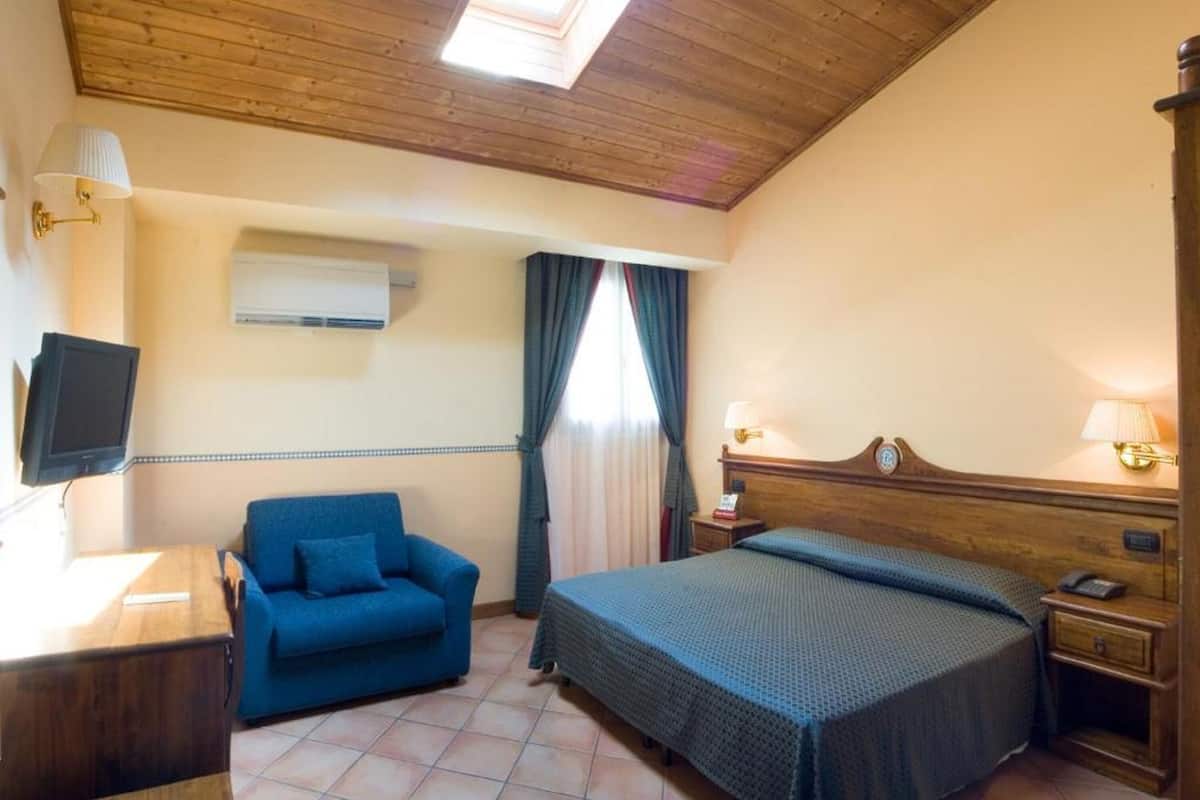 A hotel room with a wooden ceiling, a bed with a headboard, a blue sofa, a desk with a lamp, and a television.