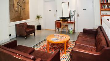 A living room with a brown leather sofa, a wooden coffee table with magazines, a wooden desk with a chair, and a bookshelf.