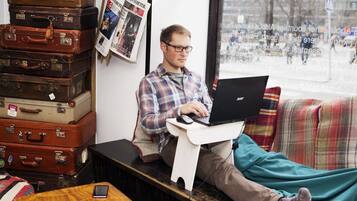 Een persoon zit op een bank met een laptop, omringd door stapels koffers, in een kamer met een raam en kranten aan de muur.