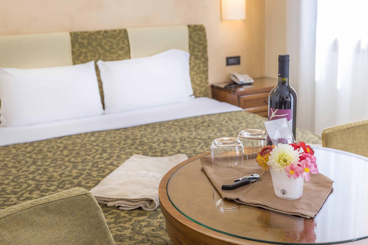 A hotel room with a bed, a glass table with a bottle of wine, a vase with flowers, and a remote control.