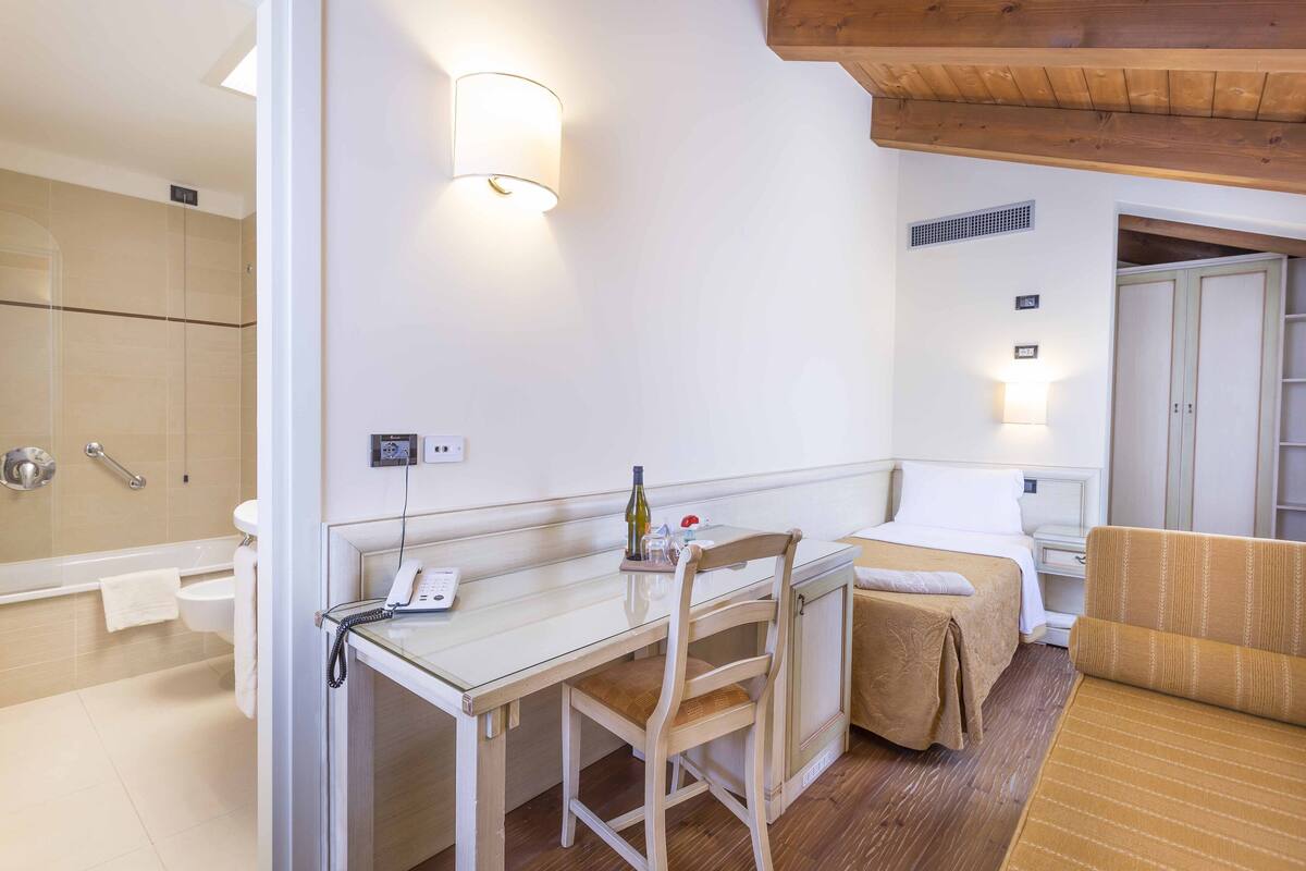 A hotel room with a glass desk, a chair, a bed with a brown cover, a wooden floor, and a white wall with a light fixture.