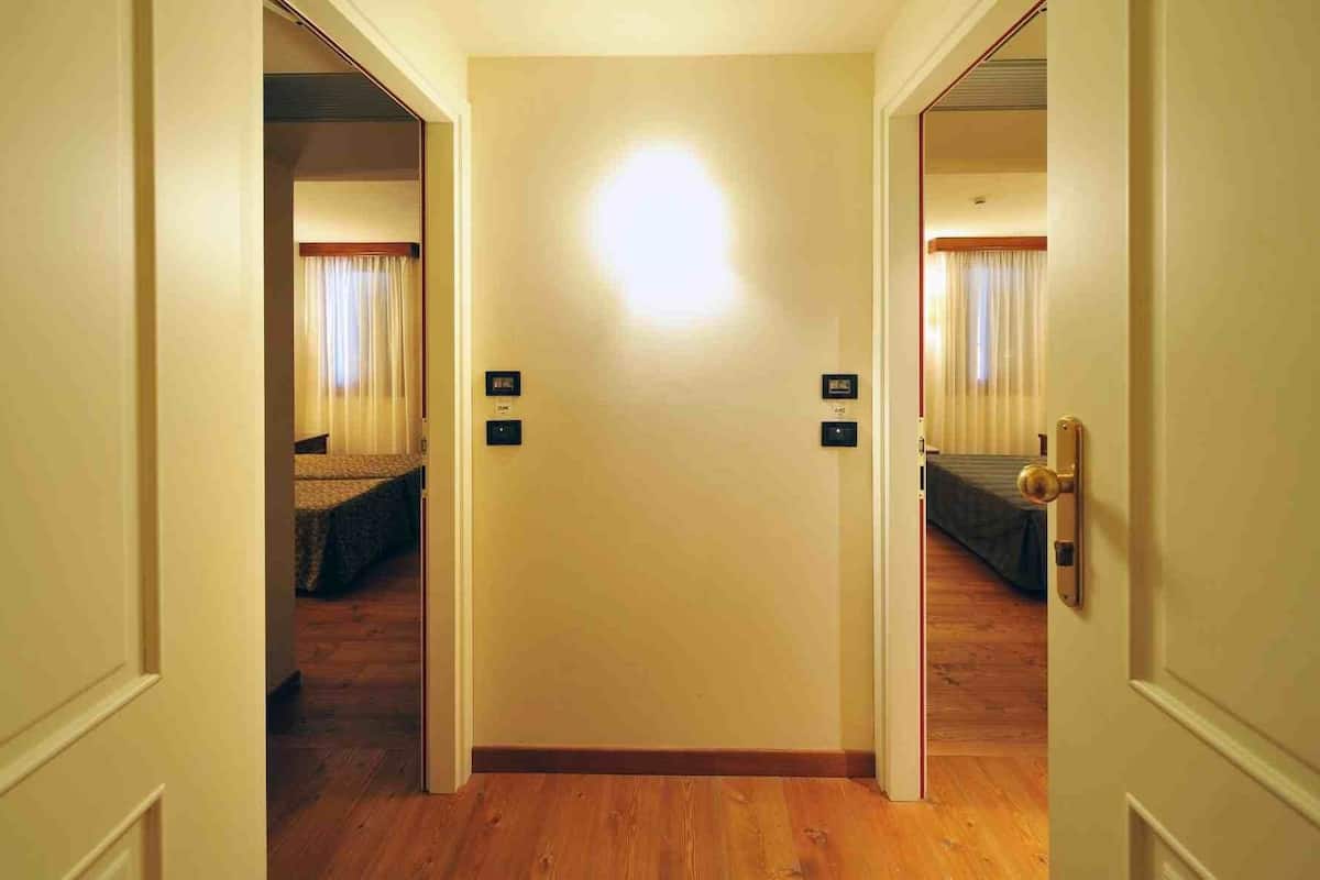 A hotel room with a wooden floor, a bed with a dark cover, and a door with a light switch and a lock.