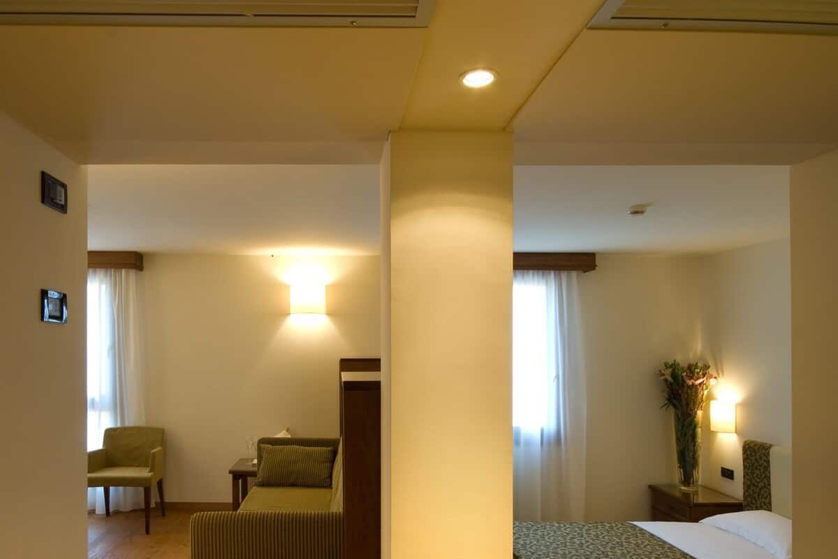 A hotel room with a bed, a chair, a small table, and a nightstand with a lamp.
