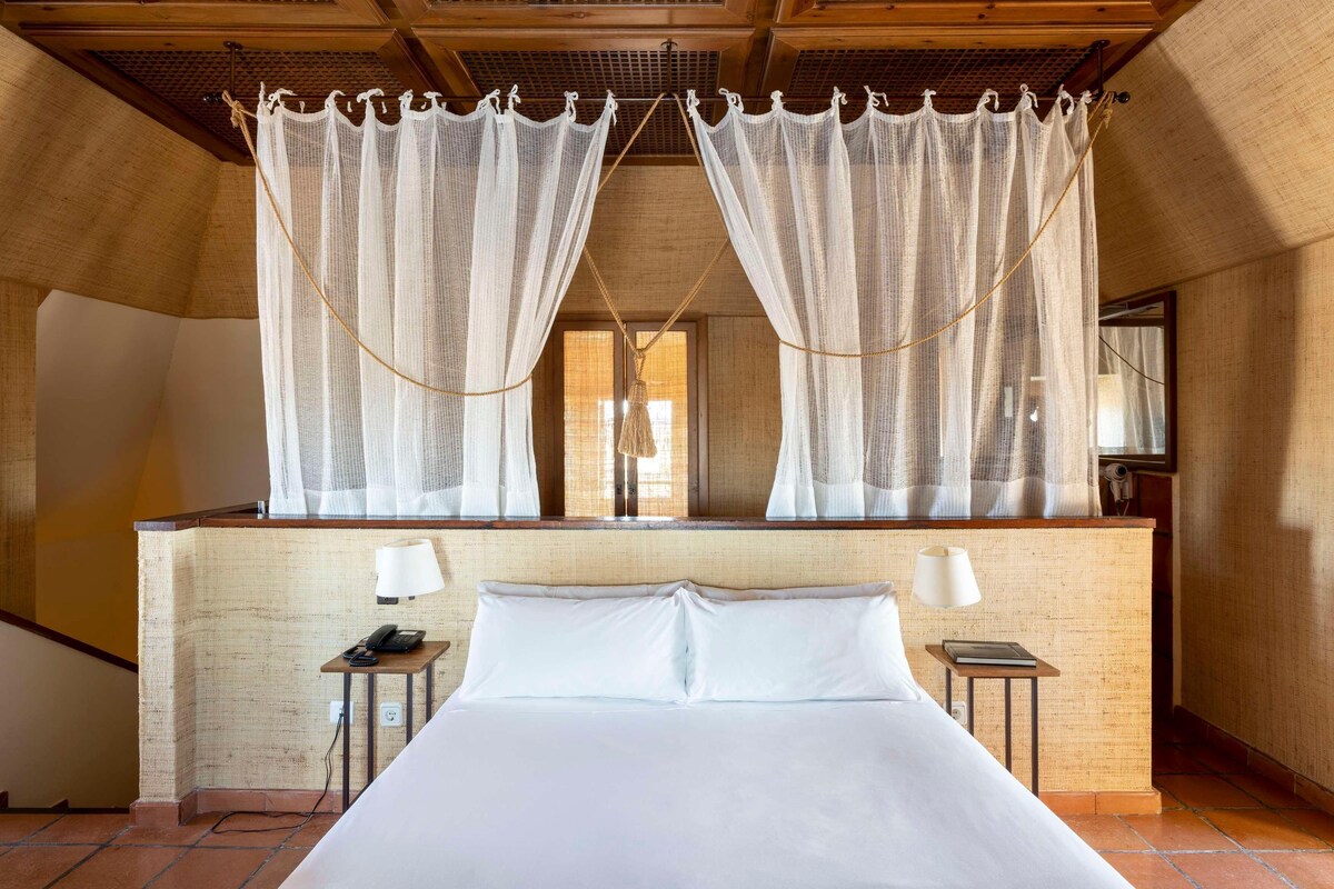 A bedroom with a large bed, two bedside tables, a nightstand, and a window with sheer curtains.