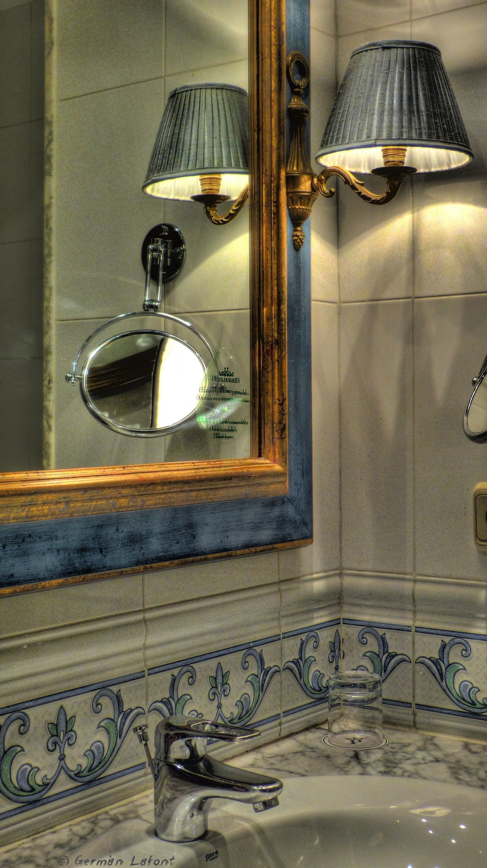 Un baño con un espejo grande, dos lámparas de pared, un lavamanos con grifo y azulejos decorativos.