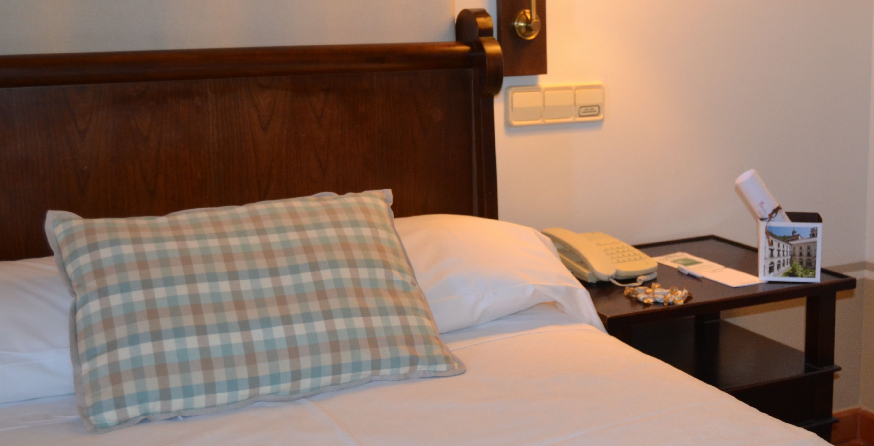 A neatly made bed with a checkered pillow, a bedside table with a telephone and a card holder, and a wall-mounted light switch.