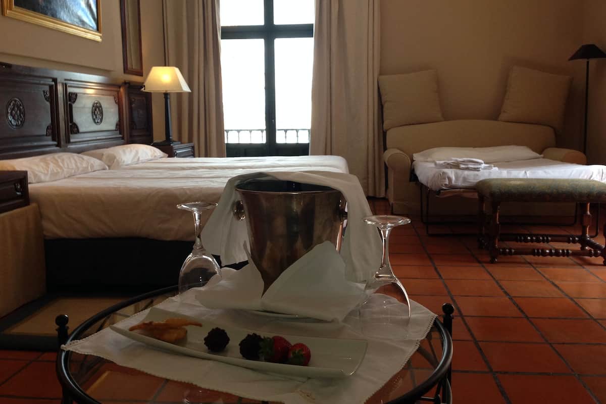 Hotelkamer met een bed, een sofa, een eettafel met eten en drinken, en een groot raam met gordijnen.