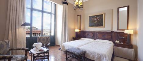Habitación de hotel con una cama grande, dos mesitas de noche, una silla, una pequeña mesa con tetera y taza, un cuadro en la pared y una ventana con cortinas.
