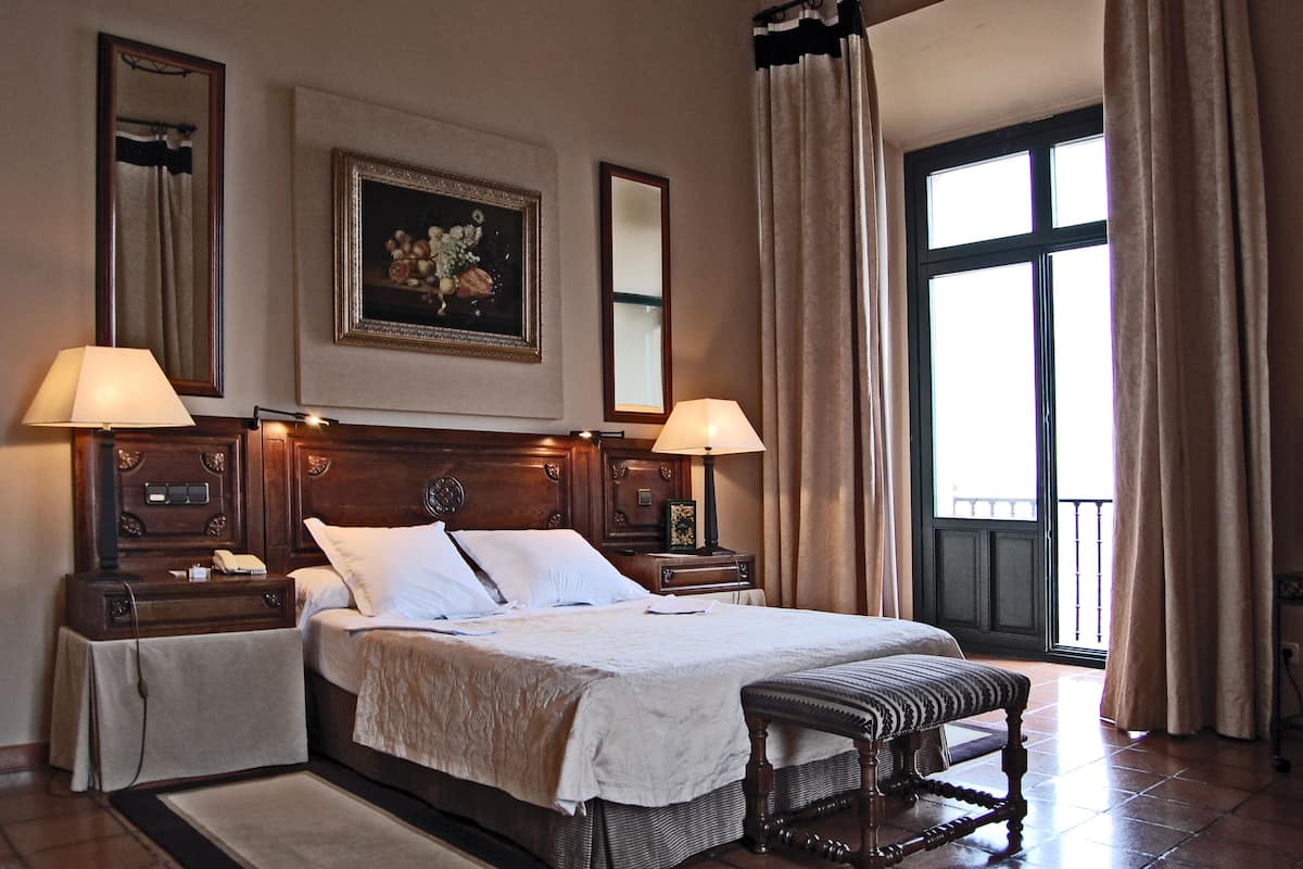 Een hotelkamer met een groot bed, twee nachtkastjes, een bankje, een schilderij en een raam met gordijnen.